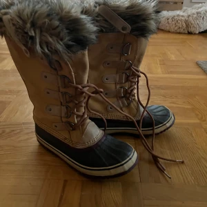 Sorel vinterboots med fuskpäls - Snygga vinterboots från Sorel i beige mocka med bruna detaljer och svart gummisula. Skorna har snörning framtill, högt skaft och fuskpäls runt kanten för extra värme. Perfekta för kalla dagar och riktigt sköna att ha på sig.