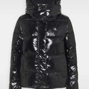 Svart glansig pufferjacka från Calvin Klein - Säljer en svart glansig pufferjacka från Calvin Klein med hög krage och huva. Jackan har dragkedja och tryckknappar framtill samt diskret CK-logo på bröstet. Perfekt för kalla dagar och ger en cool vibe till din outfit.