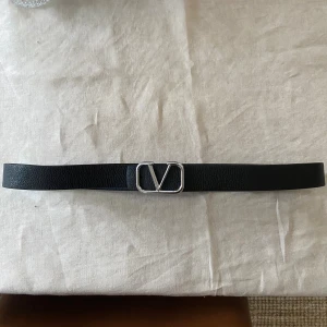 Svart skärp från Valentino - Snyggt svart skärp från Valentino med klassiskt V-spänne i silverfärgad metall. Bältet är gjort i äkta skinn med en stilren och enkel design som passar till många outfits. Perfekt accessoar för att lyfta din look.