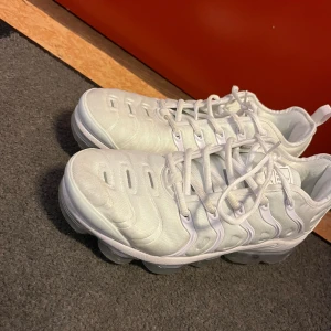 Nike Air Vapormax vita sneakers - Säljer ett par fräscha Nike Air Vapormax sneakers i helvitt med genomskinlig sula. Skorna har en futuristisk design med tydliga detaljer och snörning. Ovandelen är i syntetmaterial och mesh för en sportig vibe. Perfekta för dig som vill sticka ut med en clean look.