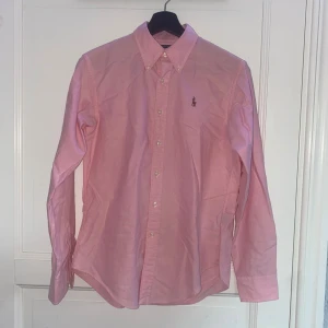 Rosa skjorta från Ralph Lauren - Snygg rosa skjorta från Ralph Lauren i klassisk passform. Skjortan har knappar framtill, lång ärm och ikonisk broderad logga med polospelare på bröstet. Tillverkad i mjuk bomull som känns skön mot huden. Perfekt för dig som gillar stilrena och tidlösa plagg.