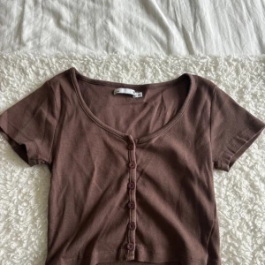 Brun ribbad topp med knappar - Kortärmad brun topp i ribbat bomullstyg med rundad urringning och knappar framtill. Croppad modell.