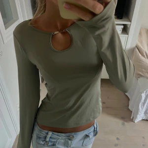 Olivgrön långärmad topp med cut-out - Säljer en olivgrön långärmad topp med rund halsringning och snygg cut-out detalj framtill som hålls ihop med en metallspänne. Modellen är croppad och har en slim passform. Perfekt att styla med jeans eller kjol för en trendig look.