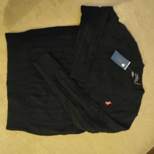 Svart kabelstickad tröja Polo Ralph Lauren - Svart kabelstickad tröja från Polo Ralph Lauren med klassisk röd broderad logga på bröstet. Tröjan har rund halsringning och långa ärmar. Perfekt för dig som gillar stilren och tidlös design.