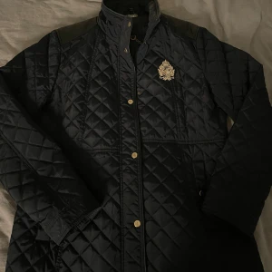 svart jacka från Ralph Lauren - Snygg quiltad svart jacka från Ralph Lauren med guldfärgade knappar och broderad logga på bröstet. Jackan har lång ärm, hög krage och ett lyxigt mönstrat innerfoder med kedjor och remmar. Perfekt för dig som gillar klassisk och stilren look. Säljer pga den inte används längre, skriv vid frågor eller intresse🥰