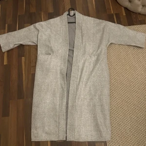 Oversized långkappa från frankrike - Säljer en stilren ljusgrå kappa i lång modell med stora fickor och öppen front. Jackan har en rak passform och är tillverkad i ett mjukt material som känns skönt mot huden. Perfekt för dig som gillar minimalistisk och clean stil.