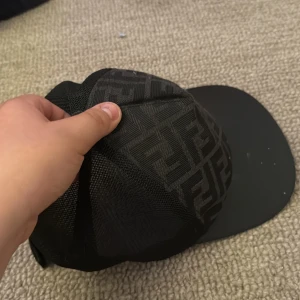 Svart keps från Fendi med mesh - Snygg svart keps från Fendi med klassiskt FF-mönster på framsidan och meshmaterial på sidorna och baksidan. Justerbar rem bak med gul Fendi-logga. Perfekt accessoar för en trendig look.