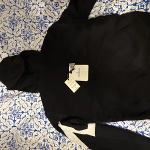 Svart cardigan med luva från Moncler - Hej, säljer min Svart Moncler cardigan med stickade svarta ärmar och vit M-mönster på ena ärmen. Cardigan har luva med snörning, dragkedja framtill och Moncler-logga på ärmen. Materialmix av dun och stickad textil ger en cool och sportig vibe.