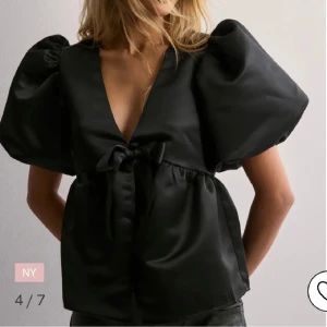 Svart blus med puffärm och rosett - Säljer en svart blus med stora puffärmar och djup v-ringning. Blusen har en söt rosettdetalj framtill och är tillverkad i ett glansigt satinliknande material. Strl s i nyskick. Nypris 500