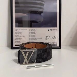 L och V rutig svart skinnbälte - Säljer ett L och V bälte med klassiskt rutigt mönster i svart och grått. Bältet har en stor, silverfärgad L och V-spänne och är tillverkat i skinn med ljusbrun insida. Perfekt accessoar för att lyfta din outfit med lyxig känsla.