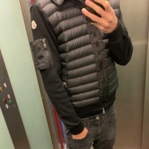 Svart cardigan med huva från Moncler - Snygg svart cardigan från Moncler med stickade svarta ärmar och vadderad kropp. Västen har huva, dragkedja framtill, fickor med tryckknapp och en liten ärmficka med dragkedja. Klassisk Moncler-logga på ärmen, perfekt till hösten!