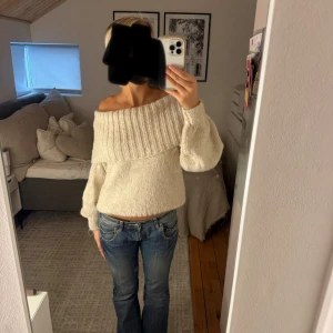 Offshoulder stickad tröja i beige/crèmefärg - Supermysig offshoulder stickad tröja i beige/crèmefärg med bred ribbad kant runt axlarna. Tröjan har långärmade puffärmar och en loose passform som ger en chill vibe. Perfekt att styla med jeans eller kjol för en trendig look.