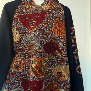Kenzo hoodie  - Unik hoodie från Kenzo med broderade motiv av vargar, tigrar och björnar i rött, orange, grått och beige. Svarta ärmar med Kenzo Paris-broderi i färgglada toner. Tröjan har huva och långärmad design, perfekt för dig som gillar statement-plagg.