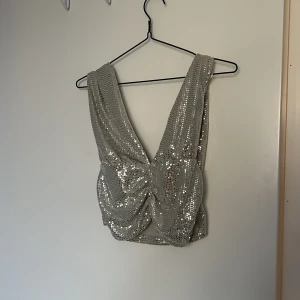 Silvrig paljettopp från Gina Tricot - Säljer en glittrig silvrig topp från Gina Tricot med breda axelband och djup v-ringning både fram och bak. Toppen är croppad och täckt av paljetter som ger ett riktigt party-vibe. Perfekt för dig som vill sticka ut med lite extra bling.