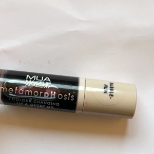MUA Metamorphosis Lip & Cheek Oil - Färgomvandlande lip & cheek oil från MUA Makeup Academy i nyansen Bubble-Hun. Oljan kommer i en smidig tub med svart och beige design, perfekt för att ge både läppar och kinder en fräsch glans och färgskiftning.