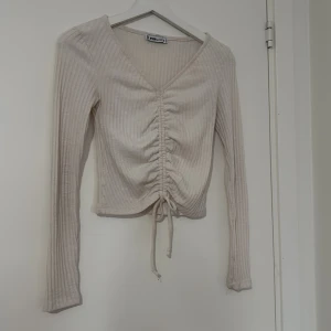 Beige ribbad omlottopp med snörning - Säljer en beige ribbad topp från Popj med lång ärm och snygg v-ringning. Toppen har snörning framtill som ger en omlott-effekt och croppad passform. Perfekt för dig som gillar stilrena detaljer och mjuka material.