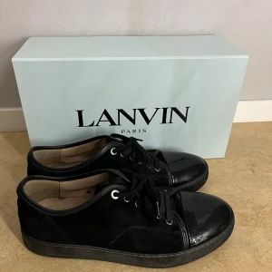 Svarta sneakers från Lanvin i mocka - Snygga svarta sneakers från Lanvin med ovandel i mocka och lackad tå. Klassisk rund tå, svarta snören och diskret logga på sulan. Skorna har en platt sula och stilren design som passar till det mesta. Levereras med originalkartong och dustbag. 