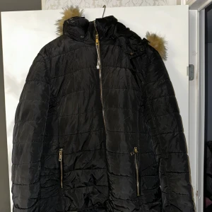 Svart pufferjacka med fuskpäls - Svart pufferjacka med huva och snygg fuskpälsdetalj i brunt. Jackan har guldiga dragkedjor på fickorna och framtill, samt quiltad design. Perfekt för kalla dagar med sitt mjuka och varma innerfoder.