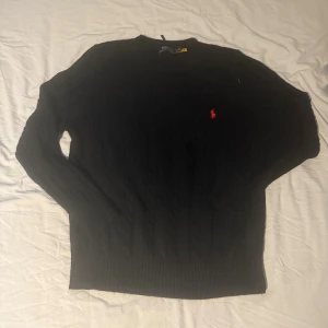 Svart stickad tröja från Polo Ralph Lauren - Svart kabelstickad tröja från Polo Ralph Lauren med klassisk röd logobroderi på bröstet. Rund halsringning och ribbade muddar vid ärmslut och nederkant. Perfekt för dig som gillar stilrena och tidlösa plagg med snygga detaljer.