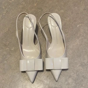 Vita pumps med rosett från Zara - Intressekoll Snygga vita pumps från Zara med spetsig tå och elegant rosett framtill. Skorna har smal klack och öppen häl med tunn rem. Tillverkade i glansigt syntetmaterial som ger en stilren look. Perfekta för dig som vill sticka ut med en chic detalj. Använd fåtal gånger, får se om jag vill sälja☺️