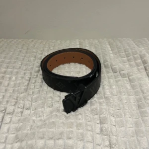 Svart skärp i skinn - Snyggt svart bälte från H&M med klassiskt monogrammönster och spänne. Bältet är gjort i skinn med en brun insida och har flera hål för justering. Perfekt accessoar för att lyfta din outfit.