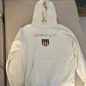 Vit hoodie från GANT med emblem - Vit hoodie från GANT i mjuk bomull med stort tryckt GANT-logga och broderat emblem med stjärnor och ränder på bröstet. Tröjan har huva med metallöljetter, känguruficka och långa ärmar. Perfekt för chill dagar och snygg till jeans.