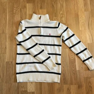 Vit randig stickad tröja Polo Ralph Lauren - Snygg vit stickad tröja från Polo Ralph Lauren med svarta horisontella ränder och röd logga på bröstet. Tröjan har half zip, hög krage och långa ärmar. Tillverkad i mjuk bomull, perfekt för dig som gillar klassisk och clean stil. Pris är diskuterbart. Fler bilder kan ges vid begäran.