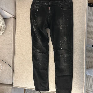 Svarta Dsquared2 jeans med slitningar - Svarta jeans från Dsquared2 med coola slitningar och vita färgstänk. Jeansen har fem fickor, smal passform och klassisk knappgylf. Snyggt tvättad finish och detaljer som ger en edgy vibe. Perfekta för dig som vill sticka ut med din stil.