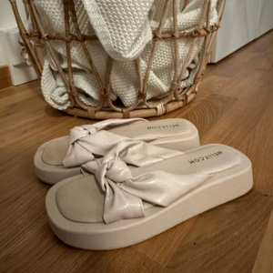 Beige sandaler med knut från Nelly - Snygga beige sandaler från Nelly med breda remmar i syntetmaterial och trendig knutdetalj upptill. De har platt sula och öppen tå, perfekt för varma dagar. Stilsäkra och lätta att matcha till sommarens outfits.
