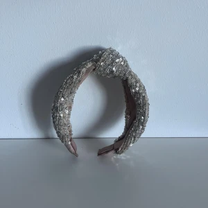 Glittrigt silverdiadem med paljetter - Snyggt diadem i silver med glittrande paljetter och en knut framtill. Perfekt för att ge din look extra bling och attityd. Diademet har en bred form och är täckt av små paljetter som reflekterar ljuset. Insidan är mjukt fodrad i beige tyg för skön komfort.