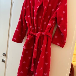 Röd fleece morgonrock med hjärtan och bälte - Mysig röd fleecejacka med rosa hjärtan, knytbälte i midjan och stora fickor. Perfekt för att chilla hemma eller mysa runt i. Långärmad och supermjuk, med en avslappnad passform och skön känsla mot huden.