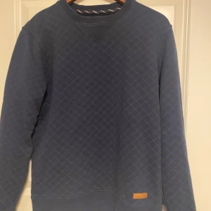 Bondelid ,Blå quiltad tröja - Säljer en snygg blå quiltad tröja från Bondelid med diskret rutmönster. Tröjan har rund halsringning, långa ärmar och en liten läderdetalj vid nederkanten. Perfekt för dig som gillar klassisk och stilren design.