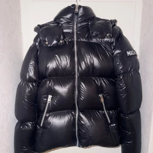 Svart pufferjacka från Mackage - Snygg svart pufferjacka från Mackage med glansig finish, hög krage och avtagbar huva. Jackan har två dragkedjeförsedda fickor framtill och tydlig logga på ärmen. Perfekt för kalla dagar när du vill hålla stilen. Storlek 38 vilket motsvarar M/L