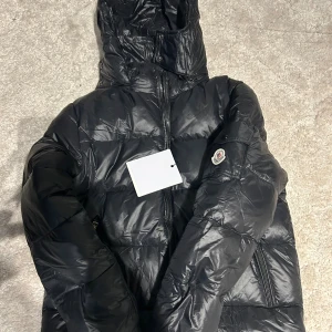 Svart Moncler pufferjacka med huva - Säljer en svart pufferjacka från Moncler med huva och dragkedja framtill. Jackan har quiltad design, lång ärm och Moncler-logga på armen. Materialet är glansigt och känns riktigt lyxigt, perfekt för kalla dagar. Snygg och stilren look som funkar till allt. Passar till M också