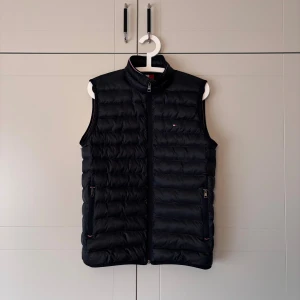 Navy blue dunväst från Tommy Hilfiger - Navy blue dunväst från Tommy Hilfiger med quiltad design och ståkrage. Västen har två fickor med dragkedja och en liten logotyp på bröstet. Perfekt för lager-på-lager och har en stilren look med diskreta detaljer. Ej använd , ny pris 2200. 