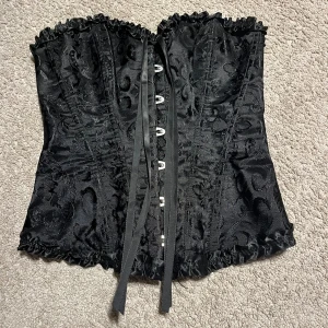Svart korsett med snörning  - Säljer en svart korsett i storlek XS med snyggt broderat mönster och glansig finish. Toppen har snörning både fram och bak, samt volangkant upptill och nedtill för extra detalj. 