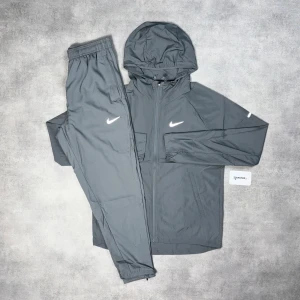  Nike Miller tracksuit grey ordinarie pris = 3200 - Snygg grå träningskostym från Nike med matchande jacka och byxor. Jackan har huva, dragkedja och två fickor, medan byxorna har elastisk midja och Nike-logga på låret. Perfekt för sport eller chill. Materialet är lätt och syntetiskt.