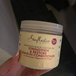 SheaMoisture Jamaican Black Castor Oil - SheaMoisture Jamaican Black Castor Oil Strengthen & Restore Leave-In Conditioner. Innehåller sheasmör, pepparmynta och äppelcidervinäger. Kommer i en rund, beige plastburk med skruvlock. Perfekt för att vårda och stärka håret. Inte använd, som ny.