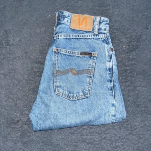 Nudie Jeans  - Snygga ljusblå jeans från Nudie Jeans med hög midja och raka ben. Klassisk femficksmodell med orange sömmar och råa, fransiga benslut för en cool look. Tillverkade i 100% bomull med Nudie-logga på bakfickan. Som det syns på bilderna är jeansen designad utan mudd längst ned och ska istället se klippta ut. Tyvärr vet jag inte vilken modell det är. Midja 33cm längd 103 cm. Som man ser på bild 4 så är det 2 små revor och på bild 5 så ser man en av bälteshålarna är sönder och ett litet hål :(