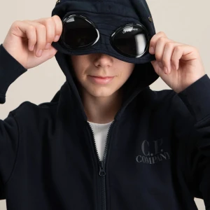 Svart hoodiejacka från C.P. Company - Svart hoodiejacka från C.P. Company med ikoniska glasögon i huvan. Jackan har dragkedja framtill, ribbade muddar och diskret logga på bröstet. Perfekt för dig som gillar streetwear och vill sticka ut med en unik detalj på huvan.