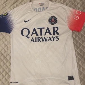 PSG vit matchtröja Nike M - Paris Saint-Germain fotbollströja i vitt från Nike med Dri-FIT material. Tröjan har PSG-emblem, Qatar Airways-tryck, blått mönster på ena ärmen och rött på den andra. Ligue 1 och GOAT-loggor på ärmarna. Perfekt för dig som älskar fotboll och PSG.
