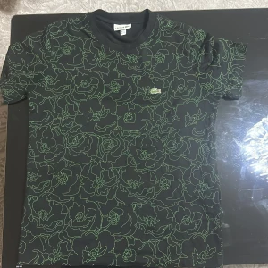 Svart blommig t-shirt från Lacoste - Svart t-shirt från Lacoste med grönt blommigt mönster över hela plagget och klassisk krokodillogga på bröstet. Skön passform och rund halsringning, perfekt för dig som vill sticka ut med en cool och unik stil.