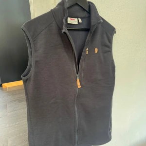 Mörkgrå fleeceväst från Fjällräven XL - Riktigt snygg mörkgrå ullväst från Fjällräven i mjuk fleece på insidan med hel dragkedja och  krage. Västen har en bröstficka med dragkedja och klassisk Fjällräven-logga på bröstet med beiga detaljer. Den är i myxket bra skick och perfekt till lager-på-lager-plagg för både uppklädda tillfällen men även till mer casual tillfällen. Den kombinerar stil och funktion väldigt bra med både ett värmande matrial som också är väldigt stilrent. Ordinarie pris var 1400