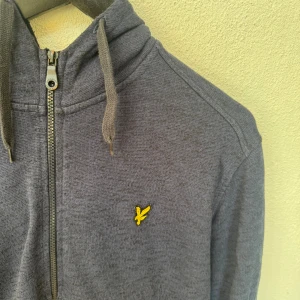 Marinblå hoodie från Lyle & Scott - Fet marinblå zip-hoodie från Lyle & Scott med dragkedja och huva med snörning. Tröjan har ett broderat gult logomärke på bröstet och två fickor framtill. Perfekt för chill dagar och enkel att matcha med jeans eller till mer uppklädda tillfällen. Tröjan är bara använd ett fåtal gånger. Ordinarie pris låg på 1000