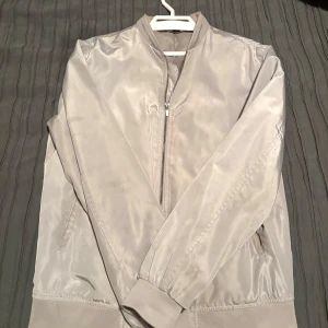 Ljusgrå bomberjacka från Zara Man - Snygg ljusgrå bomberjacka från Zara Man med klassisk ribbad krage och muddar. Jackan har dragkedja framtill och två sidofickor. Materialet är slätt och har en lätt glansig finish som ger en modern vibe. Perfekt för dig som gillar stilrena och enkla plagg.