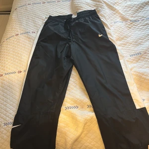 Svarta Nike täckbyxor med vita detaljer - Nike täckbyxor i svart med vita sidor och klassisk Swoosh-logga. Byxorna har elastisk midja med snörning och resår vid benslut. Lättviktigt material med meshfoder för extra komfort. Perfekta för dig som gillar sportig stil. Står ej för frakt ❤️