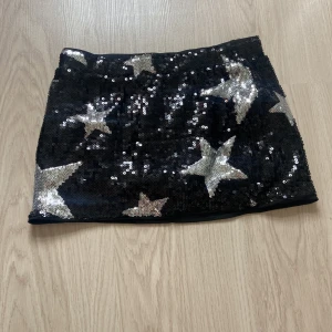Svart paljettkjol med stjärnor - Glittrig svart kortkjol täckt av paljetter och stora silvriga stjärnor. Kjolen har en tight passform och är perfekt för dig som vill sticka ut med en cool och festlig look. Ingen synlig dragkedja eller knappar.