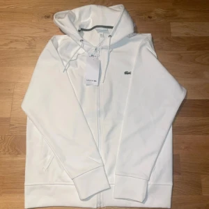 Lacoste kofta - Vit hoodie från Lacoste i klassisk passform, med huva och snörning. Tröjan har dragkedja framtill, två fickor och den ikoniska gröna krokodilloggan på bröstet. Materialet är mjuk bomull och polyesterblandning, perfekt för chill dagar.