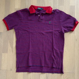 Randig röd-blå polo från Ralph Lauren - Säljer en klassisk polo t-shirt från Ralph Lauren i storlek L. T-shirten har röda och blå horisontella ränder, röd krage och ärmar samt en grön broderad logga på bröstet. Materialet är mjuk bomull och modellen har korta ärmar och knappslå vid halsen.