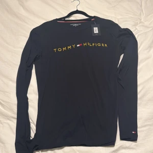 Mörkblå långärmad tröja Tommy Hilfiger, aldrig använd prislapp finns kvar - Snygg mörkblå långärmad tröja från Tommy Hilfiger med gul logotyp och det klassiska flagg-emblemet på bröstet. Tröjan har rund halsringning och är tillverkad i mjuk bomull, perfekt för en stilren och avslappnad look.